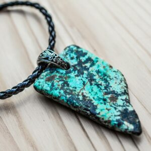 A detailed close-up of a turquoise arrowhead pendant with a braided leather chain on a wooden surface.